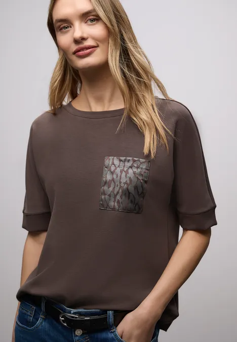 Person trägt braunes T-Shirt mit Brusttasche und Jeans, lässig stehend.